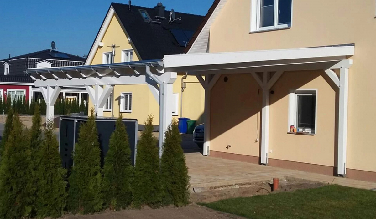 Ein weißer Holzcarport mit einem leicht abgeschrägten Pultdach grenzt an ein Wohnhaus. Der Carport verfügt über sechs Pfosten und bietet genug Platz für einen PKW.