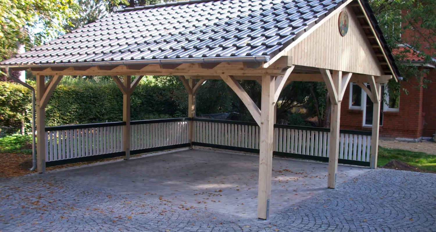 Großer Doppelcarport aus Holz. Es verfügt über ein Satteldach mit einer 30° Dachneigung und ist mit Dachsteinen gedeckt. An zwei Seiten wurde ein kleiner Zaun montiert, der sich farblich vom Carport abhebt.